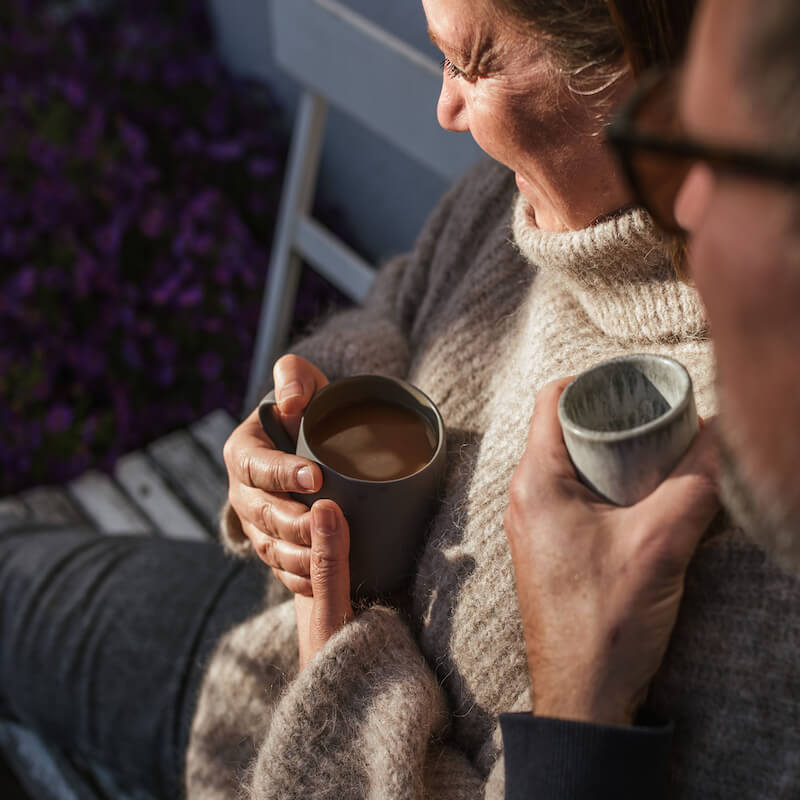 Ein Pärchen mittleren Alters sitzt mit einer Tasse langzeitgeröstetem MAYOLA Kaffee in der Sonne vor dem Haus.