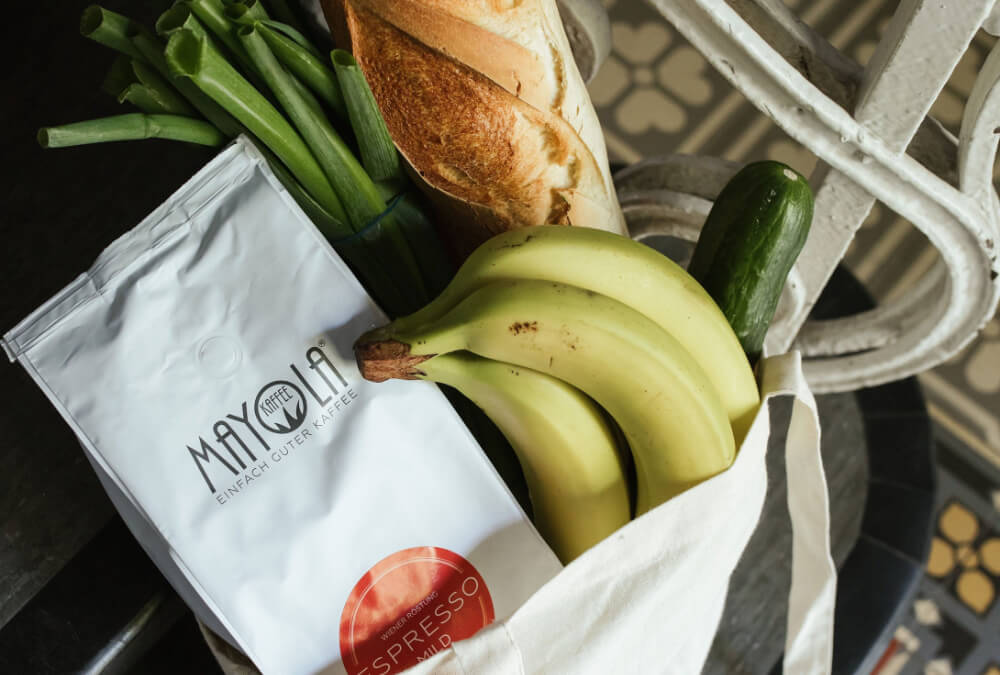 Eine Einkaufstasche aus Leinen gefüllt mit einer weißen Tüte MAYOLA Espresso Mild, Bananen, Zucchini, Frühlingszwiebeln und einem Baguettebrot. 